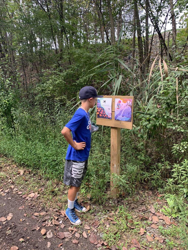 A boy about 9 years old wearing a blue baseball cap, blue shirt and gray shorts stands with his hand on his hip. He is reading a page of a book that is on a wooden stand. He is on a trail and there are trees around him.