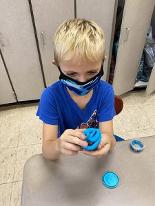 A boy with blonde hair, wearing a blue shirt and mask, holds up a round of blue play-doh in a smiley face shape.