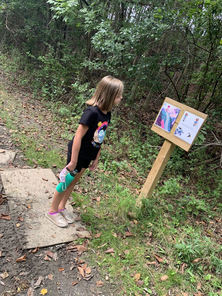 An elementary age student wearing black shorts and black t shirt holding a blue water bottle and having shoulderlength hair, reads a page from a book that is on a wooden stand. She is on a path with trees all around her.