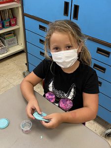 A girl with light, long hair wearing a black t shirt and white mask, holds a flattened round of blue play-doh.