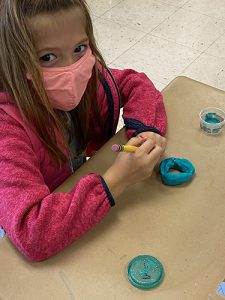 A girl with long dark blonde hair, wearing a pink jacket and pink mask, holds a pencil with a ring of blue play-doh on the table. She is looking at the camera.