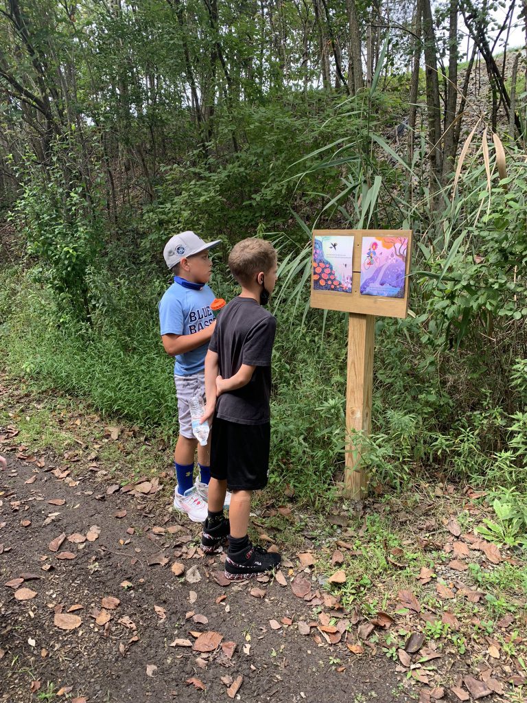 Two elementary age boys stand on a path with trees around them looking at a book open on a wooden post.