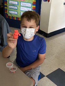 An elementary age boy with brown hair wearing a blue tshirt wearing a white mask holds up an orange man made of play-doh