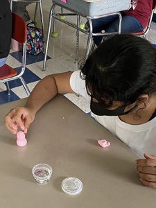 A girl with dark hair white shirt and black mask very delicately puts a bit of play-doh on a light pink figure she made.