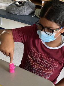 A young girl wearing glasses and a blue mask, red shirt with the word feeling repeated on the shirt, puts her pointer finger on a little pink figure made of play-doh