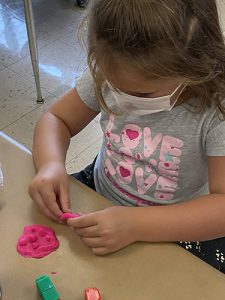 A little girl with sandy colored long hair, wearing a white mask and gray shirt that says Love Love in pink, molds pink play-doh on a table