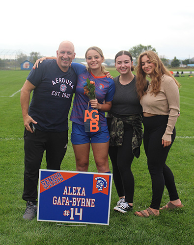 Green grass soccer field in the background and a gray sky. Four adults are standing with their arms around each other - a man on the right wearing a blue shirt and pants, a young woman in a soccer uniform of blue and orange, another young woman dressed n black and a young woman on the end with a tan sweater and black pants. All are smiling. There is a sign in front that says Alexa Gafa-Byrne #14.