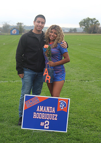 Green grass soccer field in the background and a gray sky. Two people are standing with their arms around each other, a man on the left wearing a black fleece and blue jeans, and a young woman with long blonde hair wearing a blue and orange soccer uniform. There is a sign in front that says Amanda Rodriguez.