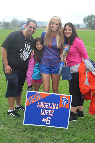 Green grass soccer field in the background and a gray sky. There is a man on the left in a black shirt, a younger girlwith long dark hair and pink jacket, a young woman with long blonde hair wearing a soccer uniform in blue and orange, and a woman on the right wearing a pink sweater and has medium length dark hair. There is a sign in front saying Angelina Lopez #5