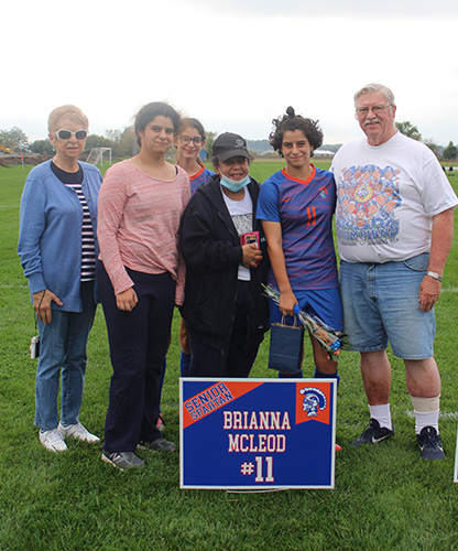Six people stand together smiling, all adults. The young woman second from right is a soccer player holding flowers and wearing a soccer uniform in blue and orange. In front there is a blue and orange sign that says Brianna McLeod #11