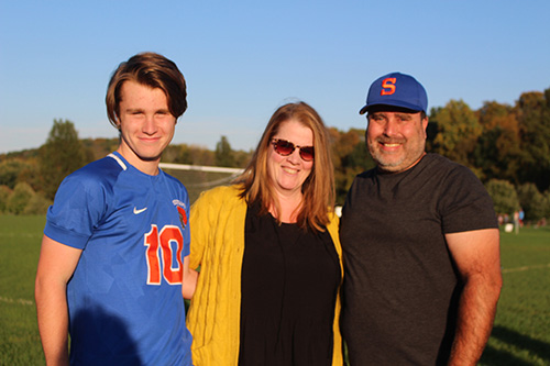 A young man wearing a blue and orange soccer uniform smiles. He has short brown hair. He is standing with a woman in the center, wearing a black shirt and gold sweater, shoulder length light hair and sunglasses, and a man on the right with a black shirt and blue cap with the letter S on it in orange. They are all locking arms.