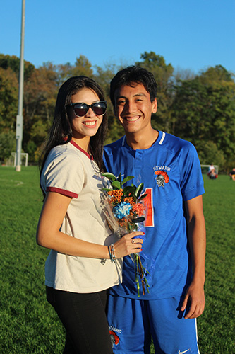 A woman on the left wearing sunglasses and a tan shirt, has long dark hair and is holding flowers. She is standing with a young man wearing a blue and orange soccer uniform. Both are smiling.