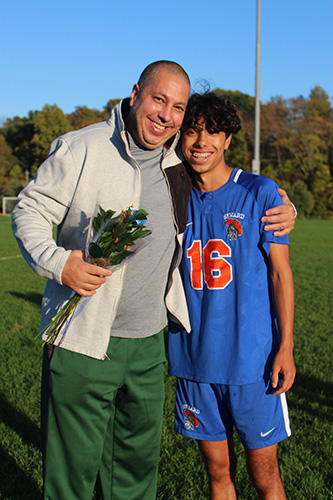 A man holding flowers stands on the left, wearing green pants and a gray shirt and jacket. He has his arm around a high school boy with dark hair smiling, wearing a blue and orange soccer uniform.