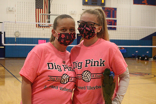 A woman with her long hair pulled back in a ponytail, wearing a pink tshirt that says Dig Pink on it, has her arm around a high school senior wearing the same design on her shirt, glasses and her blonde hair pulled up in a ponytail. They are both wearing black masks with pink ribbons printed on them.