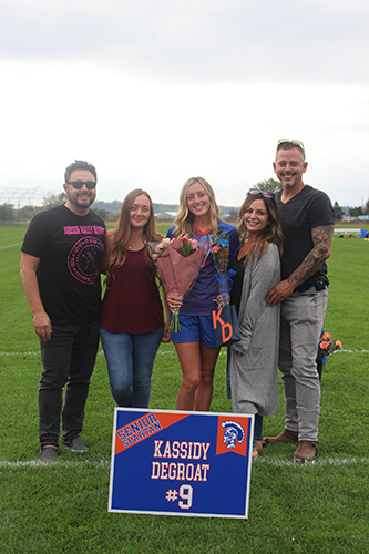 Green grass soccer field in the background and a gray sky. There are five people in front with arms around each other. In the center is a young woman with long blonde hair holding flowers and wearing a blue and orange soccer uniform. There is a sign in front that says Kassidy DeGroat