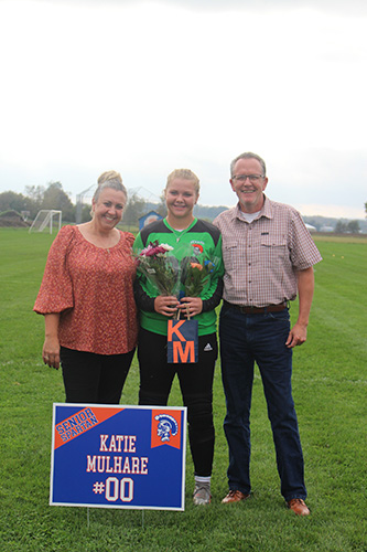 Green grass soccer field in the background and a gray sky. Three people in the photo. A woman on the left with her hair up wearing an orange shirt and black pants, a man onthe right with a short-sleeve button down shirt and black pants. In the center is a young woman, hair pulled back, wearing a green and black goalie jersey. She is holding flowers and there is a sign in front that says Katie Mulhare #00.