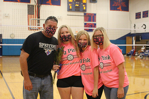 Four people are in a high school gym. There is a man on the left, and three women on the right. All of the women are wearing pink tshirts that say Dig Pink. All are wearing masks with pink breast cancer ribbons on them.