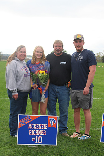 Green grass soccer field in the background and a gray sky. Four people in the photo, standing shoulder to shoulder. A woman is on the left in a gray hooded sweatshirt, Next is a young woman with shoulder-length blonde hair holding flowers, wearing a blue and orange soccer uniform, next is a man in a black shirt and jeans, on the right is a man in a blue shirt, gray shorts and wearing a baseball cap. In front is a sign that says McKenzie Richner #10