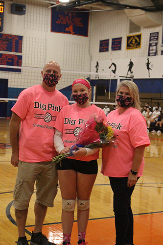 Three people in a high school gym. A man on the left, a high school girl in the center and a woman on the right. All are wearing Dig Pink t shirts. The girl in the center is holding flowers. They all have breast cancer awareness masks on.