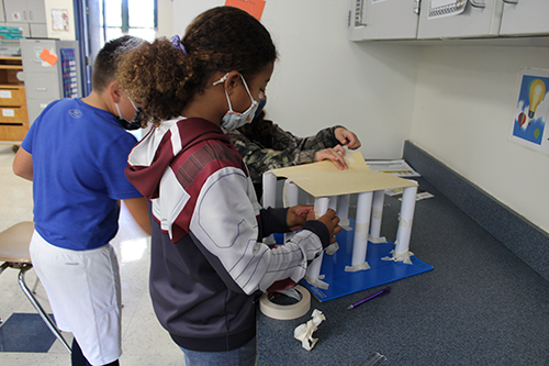 A girl wearing a ponytail, mask and red, white and blue jacket with hood puts tape on a column made of white paper. Another child places a piece of construction paper on top of it and a boy stands in the background.