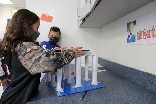 A girl wearing a mask places a large textbook on top of 10 columns made of white paper. A boy stands and watches.