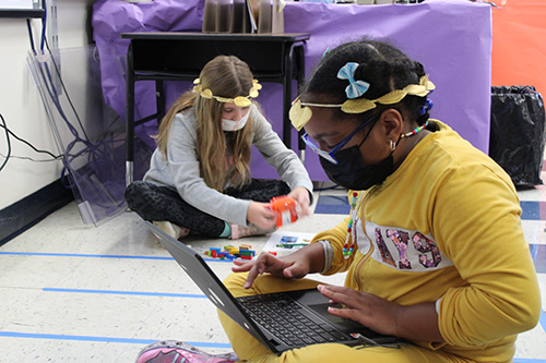 Two elementary age girls sit on the floor. Both are wearing gold leaf wreaths in their hair. One girl closest is wearing a yellow sweatsuit. She has classes and has a chromebook on her lap, that she is working on. The girl in the back has long hair and is using Legos to build a robot.