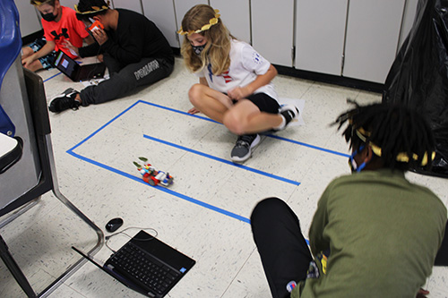 A student with long blonde hair watches as a small robot moves along the floor. Another student wearing a green shirt is watching.