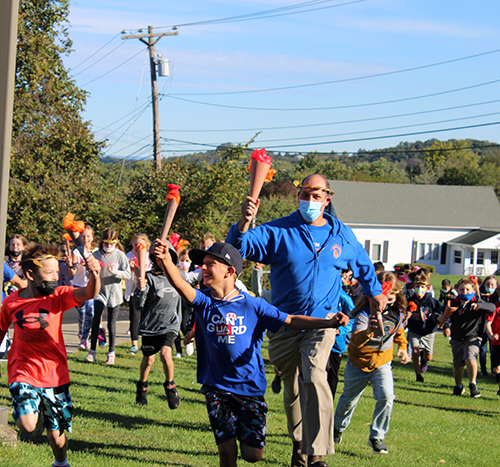 A group of elementary students and a teacher run holding torches made of cardboard tubes and orange paper.