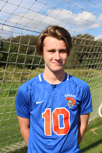 A high school boy with light hair, wearing a blue soccer jersey with the number 10 in orange and a spartan in the upper right side stands in front of a soccer net with blue skies, white fluffy clouds and trees behind him.