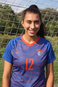 A young woman in high school with a long dark pony tail smiles. She is wearing a blue and orange soccer jersey, with the number 12 on it and a spartan on the upper right side. Behind her is blue sky, white fluffy clouds and green trees.