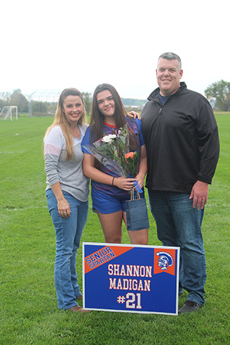 Green grass soccer field in the background and a gray sky. Three people in front - a woman on the left wearing jeans and a long-sleeve tshirt with long blonde hair, a young woman in the center with long dark hair, holding flowers, wearing a blue and orange soccer uniform, and a tall man on the right wearing a black jacket and jeans. In front is a sign that says Shannon Madigan #21.