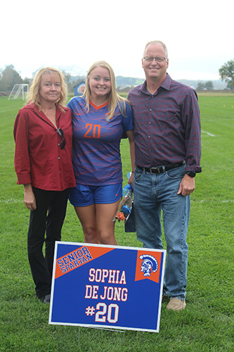 Green grass soccer field in the background and a gray sky. Three people in the photo. A woman wearing a rust-colored blouse and black pants, with shoulder-length blonde hair, a young woman with shoulder-length blonde hair wearing a blue and orange soccer uniform, and a man wearing jeans and a long-sleeve shirt. All are smiling and have arms around each other. There is a sign in front that says Sophia de Jong #20.