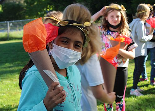An elementary scool girl wearing a blue sweatshirt, mask and a gold leaf wreath around her head, holds a torch made of cardboard tubing and orange paper.