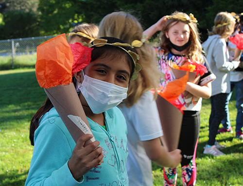 An elementary scool girl wearing a blue sweatshirt, mask and a gold leaf wreath around her head, holds a torch made of cardboard tubing and orange paper.