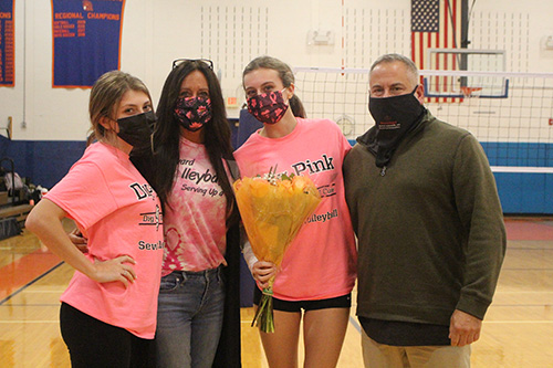 Four people in a high school gym Three women on the left have pink Dig Pink shirts on and the man on the right has a green shirt and black mask. The women also have breast cancer awareness masks on the the girl second from right is holding flowers.