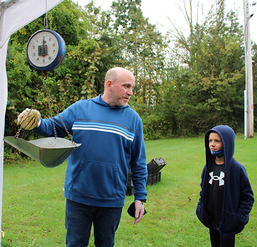 An elementary-age boy wearing a blue hoodie with a white logo on it watches as a man wearing a blue shirt with stripes across the chest puts a large vegetable on a hanging scale. There is grass and trees behind them.