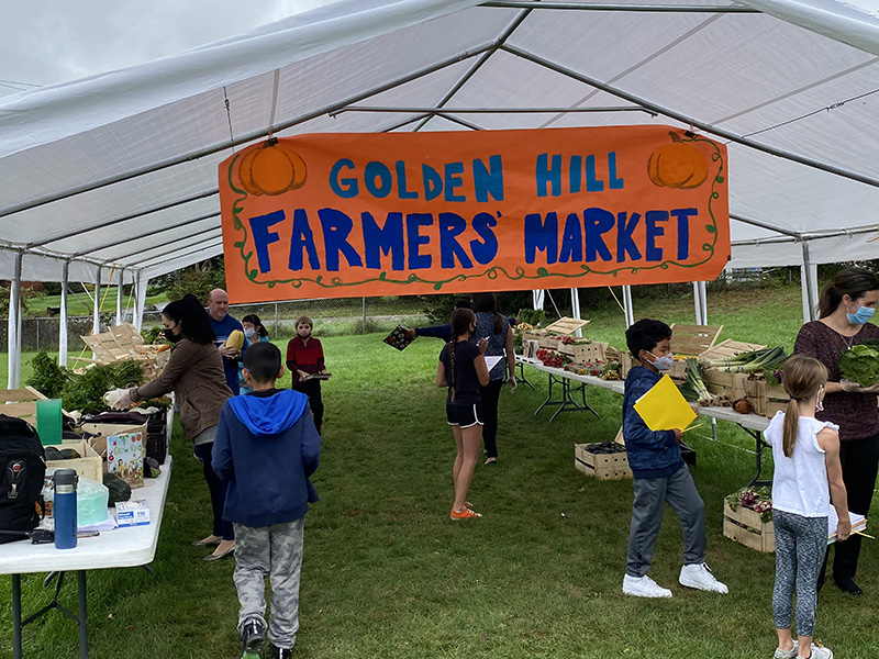 Several elementary students mull about in a tented farmers market, with bins of vegetables on tables under a large white tent. There is an orange sign that says Golden Hill Farmers Market  painted in blue hanging across the inside of the tent.