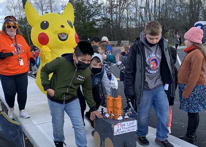 A group of three boys push a box made into a float. The boys are dressed in winter jackets and all have masks. There is a yellow Picachu balloon behind them and a woman in an orange sweatshirt