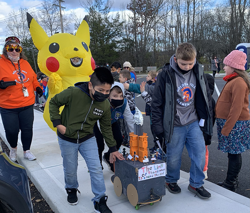 A group of three boys push a box made into a float. The boys are dressed in winter jackets and all have masks. There is a yellow Picachu balloon behind them and a woman in an orange sweatshirt