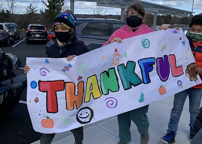 Three older elementary students carry a white banner painted in multiple colors that says Thankful. A boy in front is wearing a multi-colored knit hat and a mask, a girl in center has a pink sweatshirt, black mask, glasses, and a boy in back is wearing a green mask and a blue and orange jacket