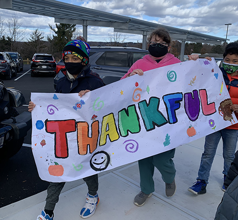 Three older elementary students carry a white banner painted in multiple colors that says Thankful. A boy in front is wearing a multi-colored knit hat and a mask, a girl in center has a pink sweatshirt, black mask, glasses, and a boy in back is wearing a green mask and a blue and orange jacket