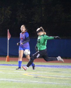 A high school girl in a green goalie's shirt and black pants kicking a soccer ball away.
