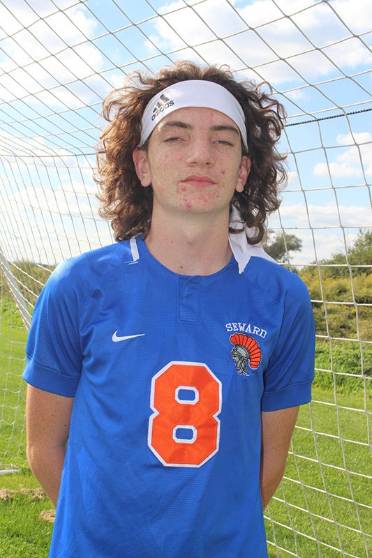 A young man with shoulder-length curly brown hair, wearing a white headband and a blue soccer jersey with the number 8 in orange. Behind him is a soccer net and blue sky and green grass and trees.