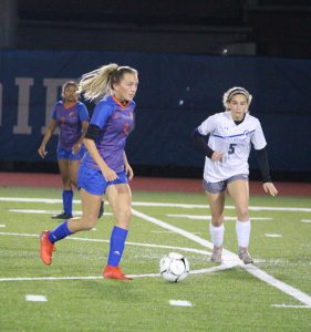 An action shot of a young woman with long blonde hair pulled back into a ponytail. She is dribbling a soccer ball on a field. She is wearing a blue soccer uniform with orange cleats. There is a girl in a white and blue uniform running toward her. This is a night game.