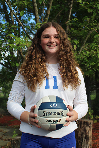 A high school gir with long curly hair smiling widely. She is holding a volleyball and wearing a white jersey with the number 11 in blue.