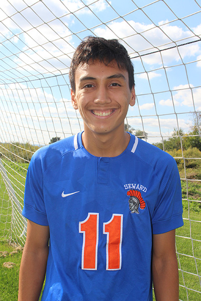 A high school boy with short dark hair smiling widely. He is standing in front of a soccer net and is wearing a blue soccer jersey with the number 11 in orange.