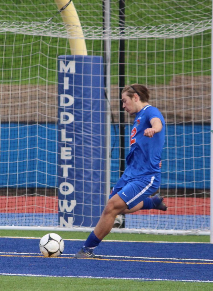 A high school boy in a blue and orange soccer uniform. He is about to kick a soccer ball away from the net.