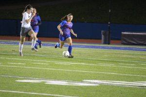 A high school girl in a blue and orange soccer uniform running on the field with a soccer ball at her foot.
