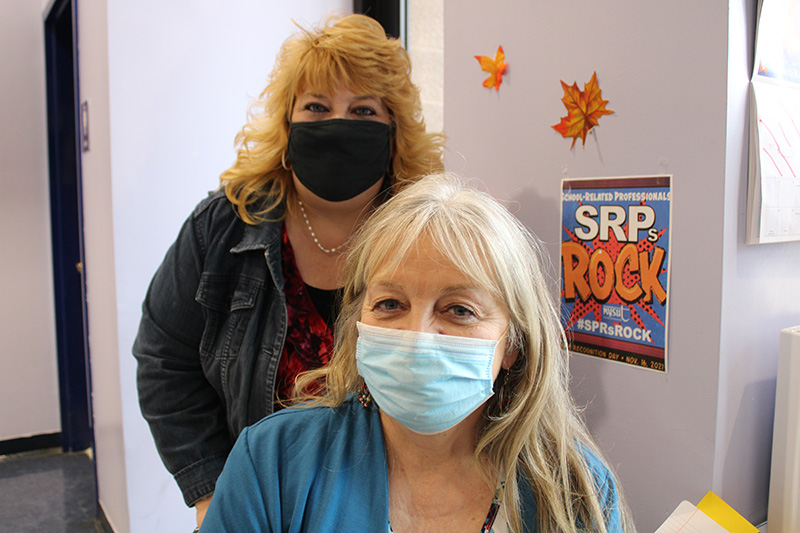 Two women. One standing in the back with shoulder-length strawberry blonde hair, wearing a denim jacket and a black mask. The women in front is sitting. She has long blonde hair and is wearing a light blue mask and blue sweater.