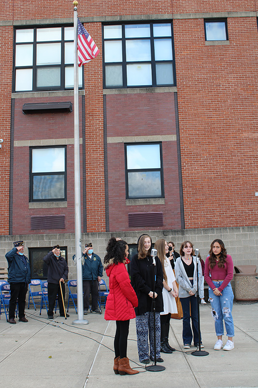 A group of five high school girls stand by two microphones. There is a brick building in the background and a flagpole with the American flag at the top. There are three older men saluting.